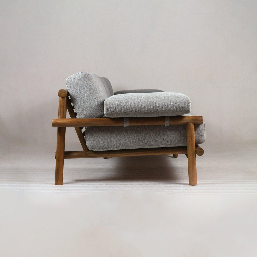 Side profile of an outdoor teak-frame sofa with warm wood finish and slatted back, featuring light gray Sunbrella fabric cushions and plush seat and back pads in a low modern coastal silhouette.