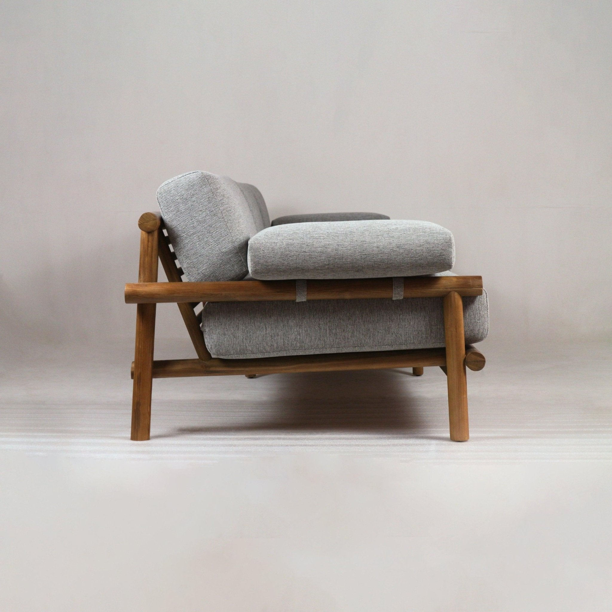Side profile of an outdoor teak-frame sofa with warm wood finish and slatted back, featuring light gray Sunbrella fabric cushions and plush seat and back pads in a low modern coastal silhouette.