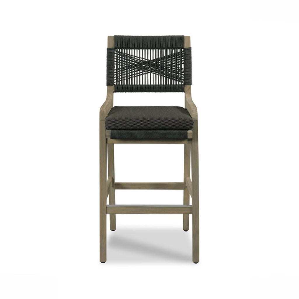 Front-facing teak outdoor bar stool with natural wood frame, dark charcoal woven rope back in a crisscross pattern and matching cushioned fabric seat, square legs and a metal footrest.