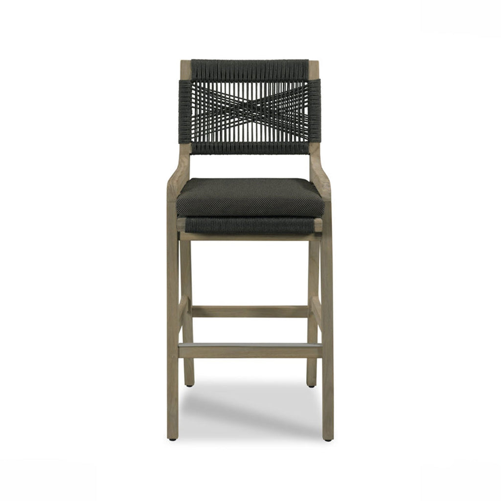 Front-facing teak outdoor bar stool with natural wood frame, dark charcoal woven rope back in a crisscross pattern and matching cushioned fabric seat, square legs and a metal footrest.