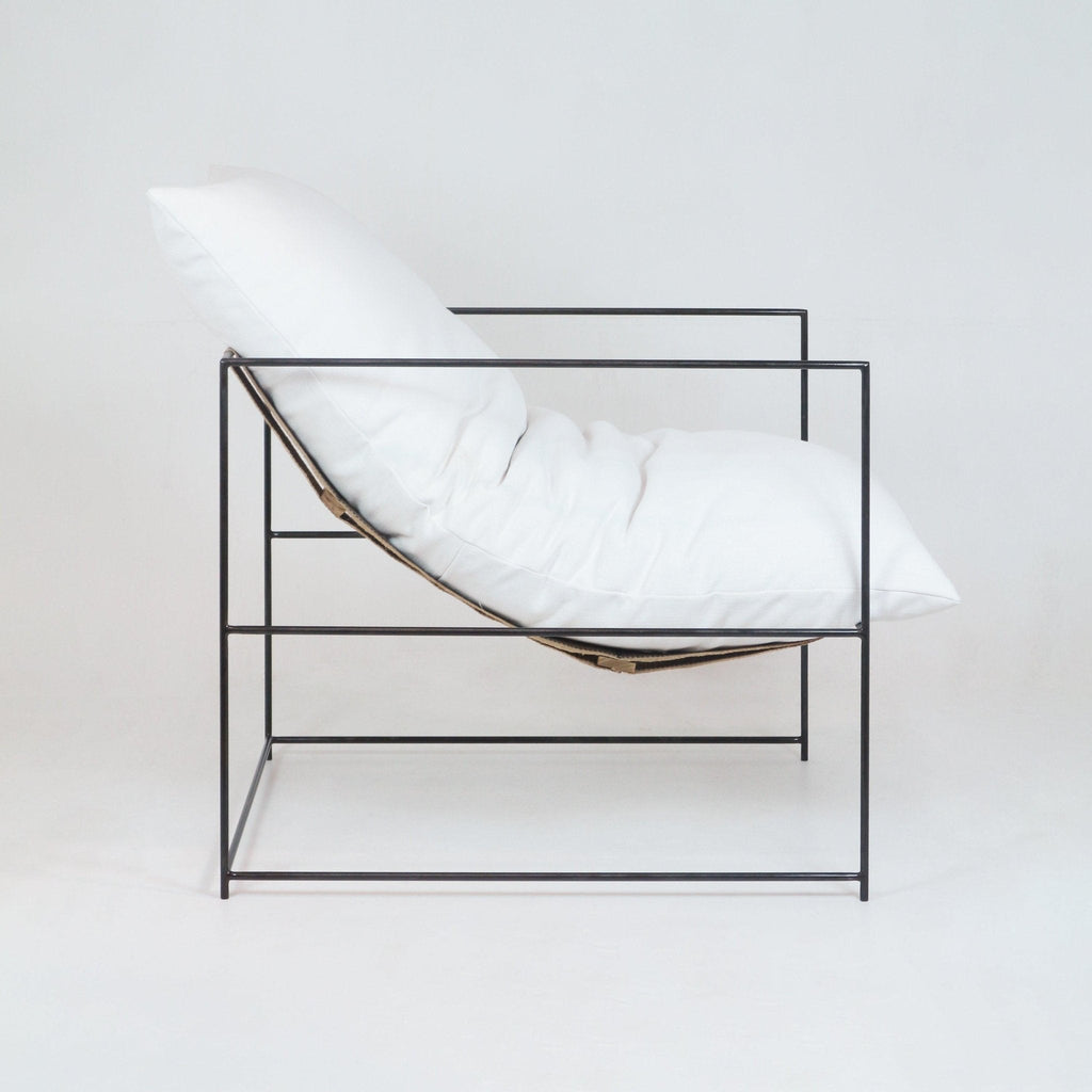 Side view of a modern pavilion chair with a plush white linen cushion and pillow suspended in a slim black metal cube frame, showcasing a minimalist contemporary accent chair for living spaces.
