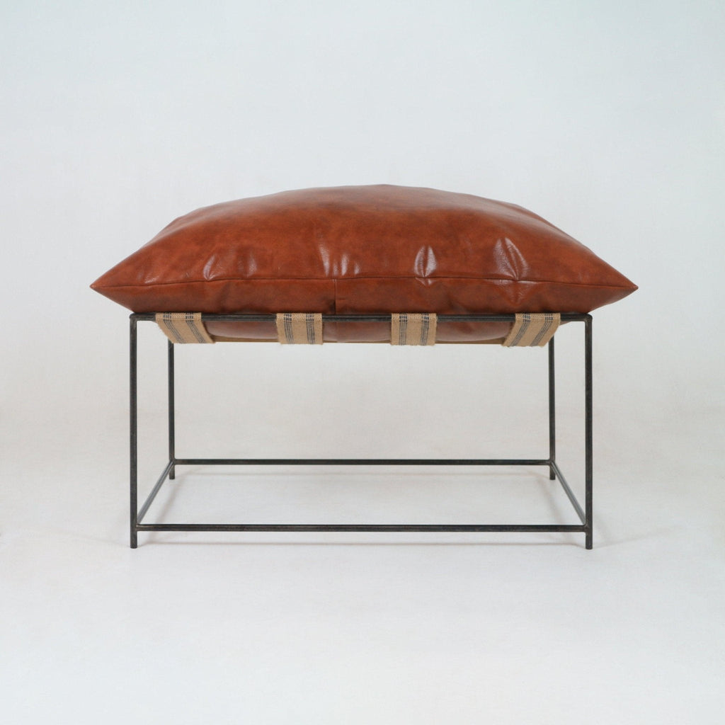 Low-profile ottoman with a plush red leather cushion resting on a minimalist black metal square frame, supported by natural woven webbing straps — a modern living room accent stool.