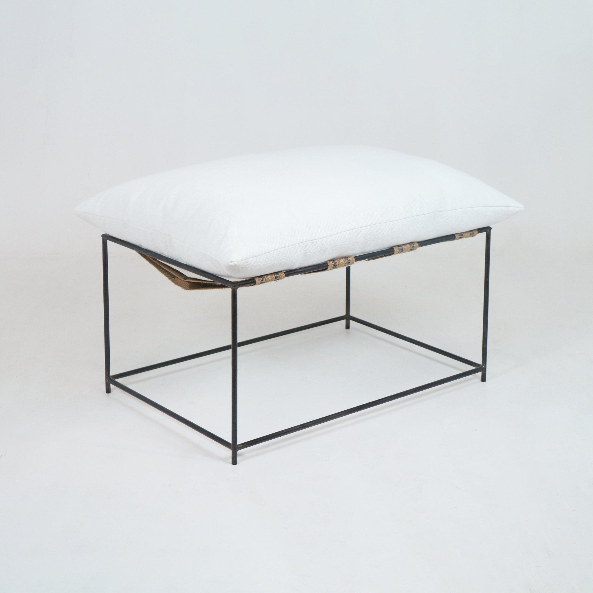 Modern outdoor ottoman with a plump white linen cushion resting on a slender black metal frame and natural woven support straps, minimalist backless design suited for patio or living spaces.