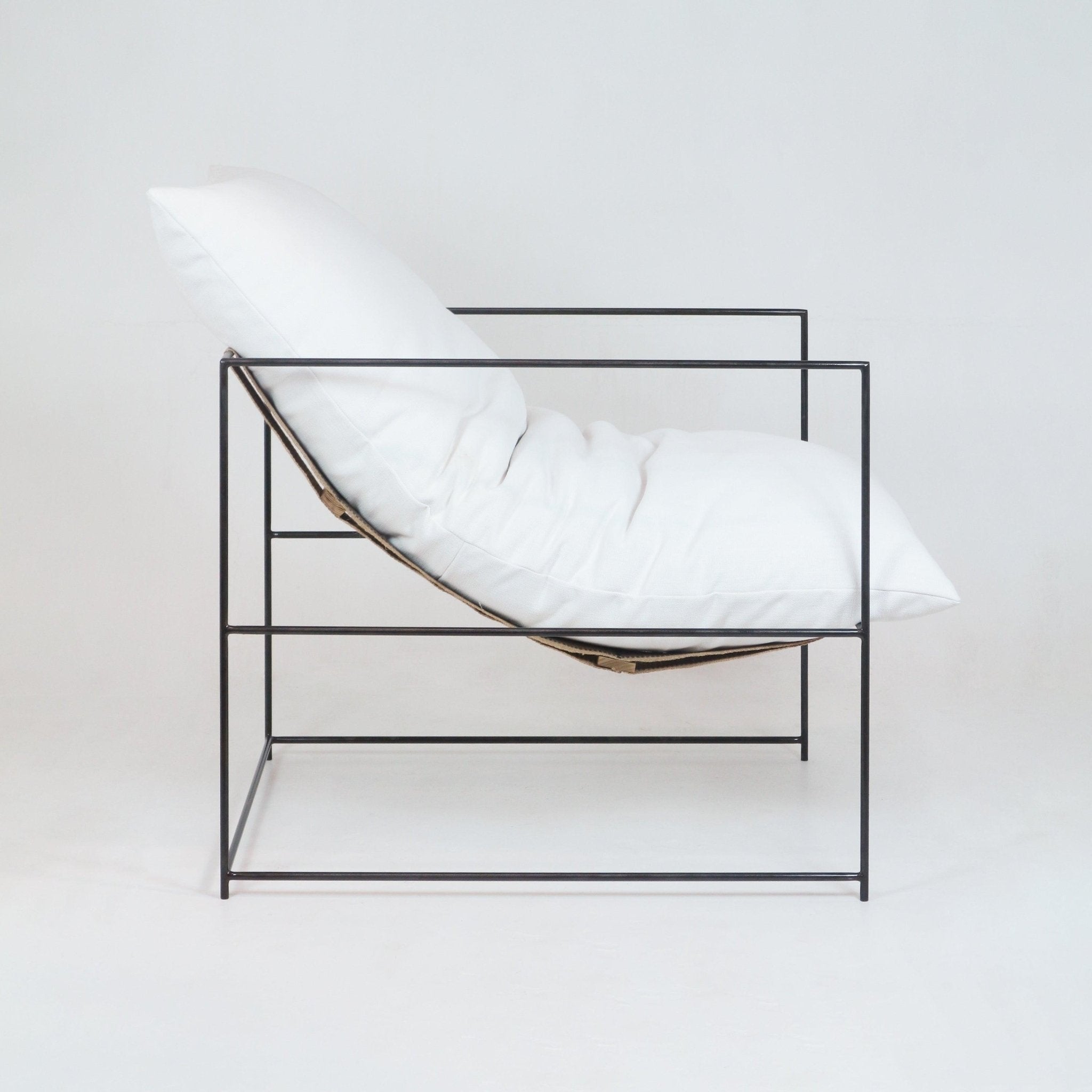 Side view of a modern pavilion chair with a plush white linen cushion and pillow suspended in a slim black metal cube frame, showcasing a minimalist contemporary accent chair for living spaces.