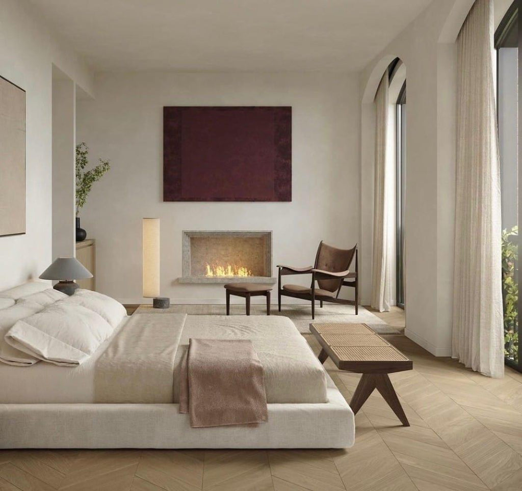 Natural wood bench with a woven Vienna cane top and splayed wooden legs, placed at the foot of a low beige upholstered bed in a minimalist modern bedroom with neutral linens, fireplace, and arched windows.