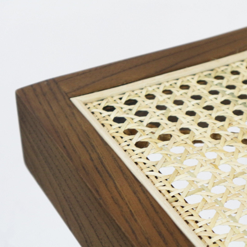 Close-up of a footstool corner showing a natural woven cane seat insert set into a medium-brown solid wood frame, highlighting the cane's hexagonal weave and smooth wood grain in a mid-century modern style.