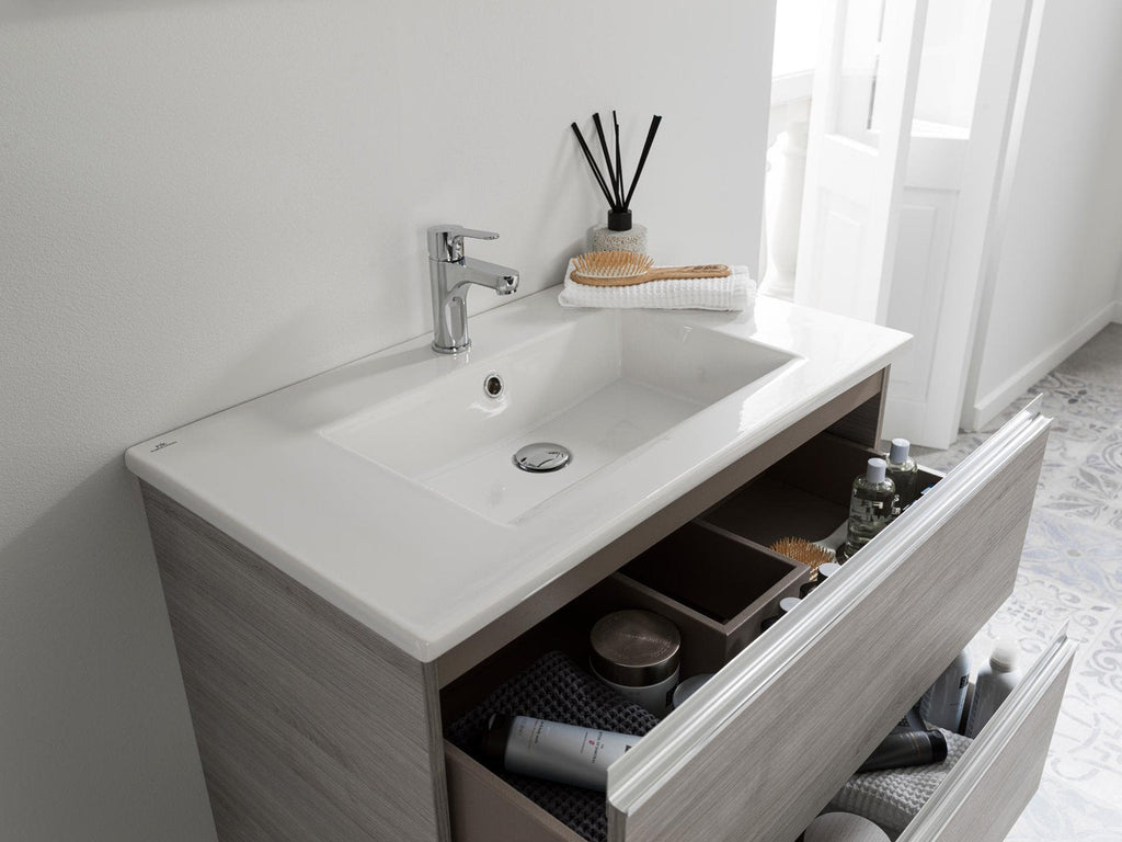Modern stone-colored solid wood single-sink vanity with a white rectangular ceramic top and chrome single-handle faucet, two open drawers showing organized storage and toiletries in a bright bathroom setting.