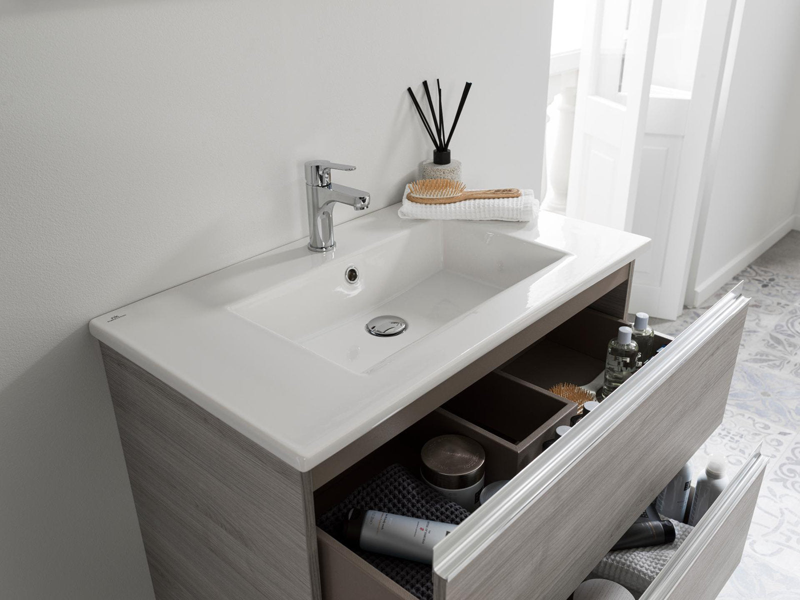 Modern stone-colored solid wood single-sink vanity with a white rectangular ceramic top and chrome single-handle faucet, two open drawers showing organized storage and toiletries in a bright bathroom setting.