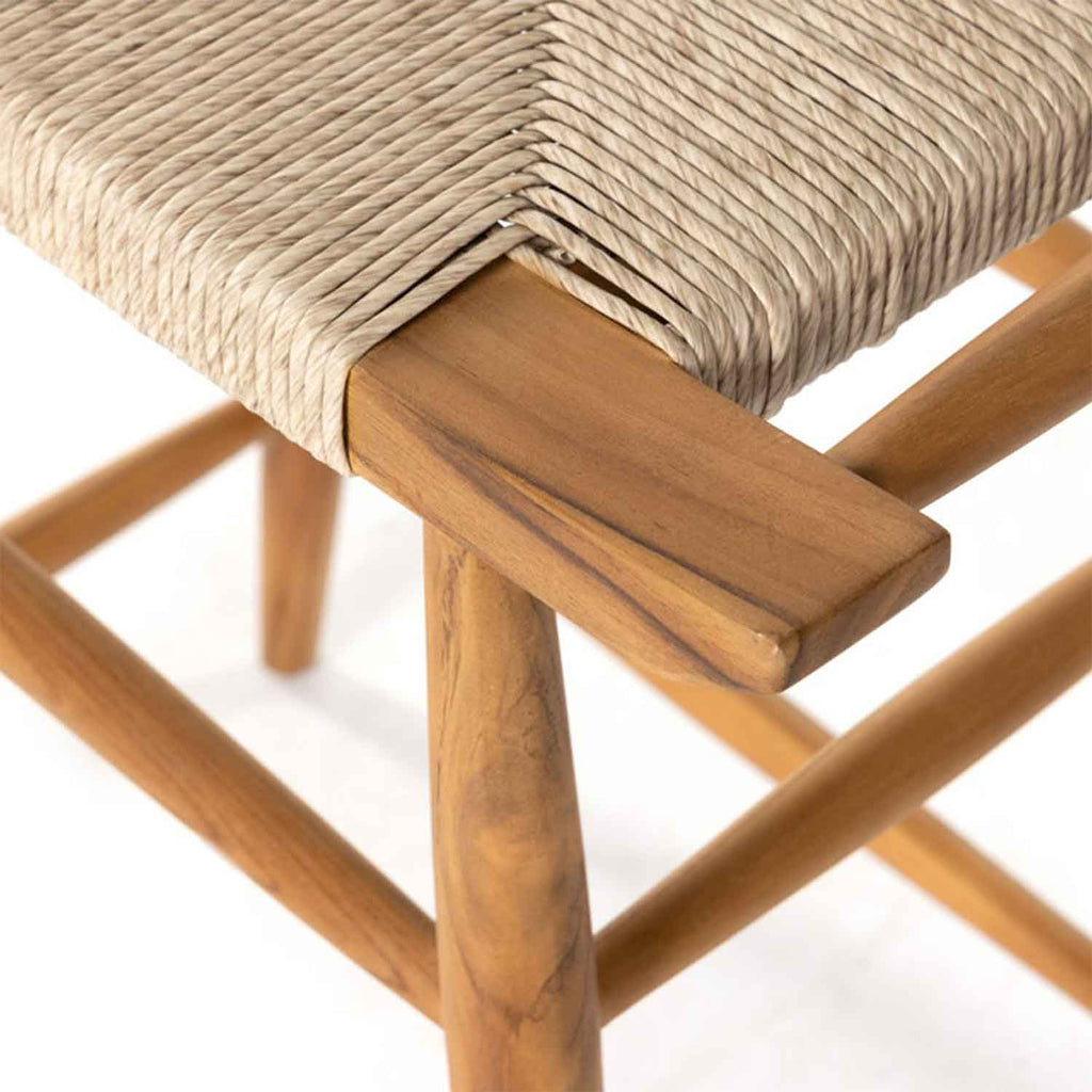 Close-up of a teak outdoor bar stool corner showing natural teak wood legs and stretcher with a tightly woven beige natural-fiber seat, highlighting wood grain and handwoven rope texture.