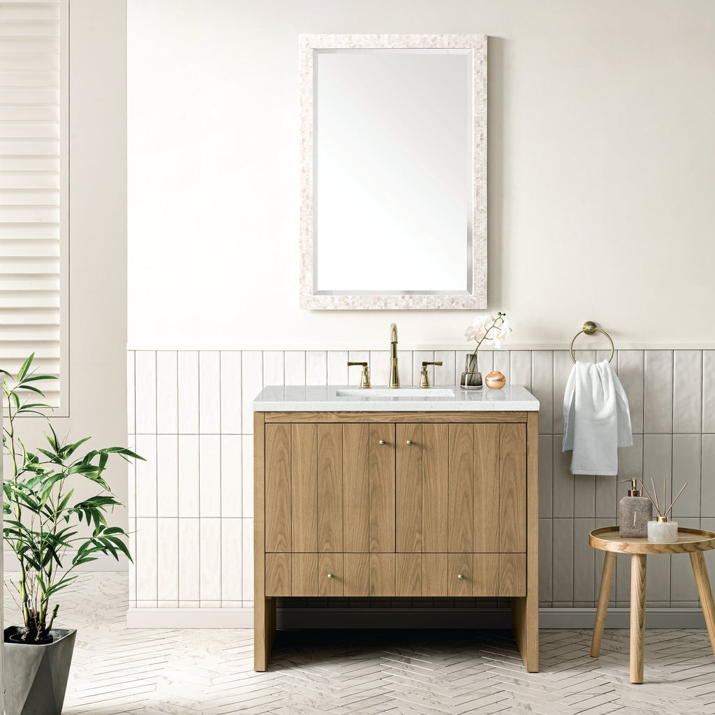 A modern bathroom features the NUEMA Sala 36 Single Bathroom Vanity with brass hardware, a stone countertop, and a rectangular mirror above. A brass faucet, towel ring, white towel, small plant, and wooden stool complete the minimalist look.