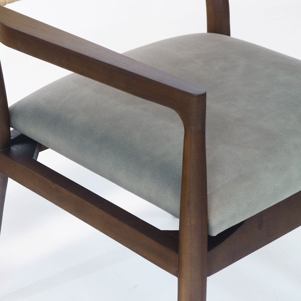 Close-up of a dining chair showing a natural wood arm and frame with a cushioned blue faux-leather seat, subtle leather texture, and a hint of the rope-wrapped backrest at the top left.
