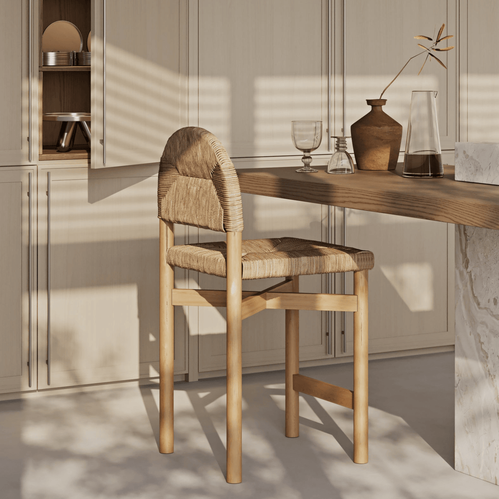 Natural teak counter stool with woven seagrass seat and rounded woven back, placed at a wood kitchen island in warm coastal tones with sunlight highlighting the wood grain and woven texture.