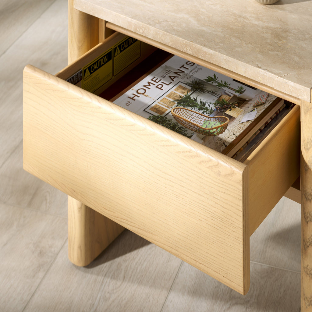 Close-up of a natural wood nightstand with an open drawer showing light oak grain, a beige marble tabletop edge above, and a lifestyle magazine tucked inside.
