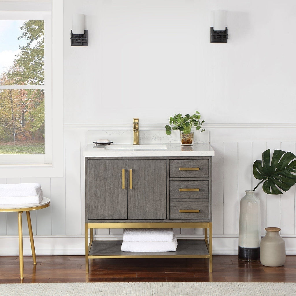 36-inch freestanding modern gray solid-wood vanity with stone countertop and white undermount sink, brass faucet, brass handles and base, two cabinet doors, three drawers and a lower shelf with folded white towels.