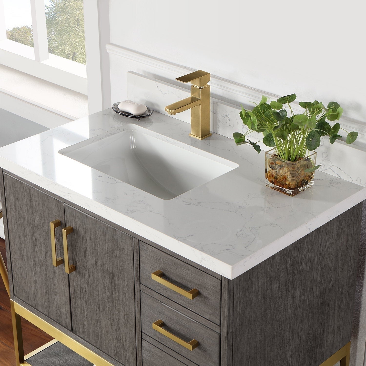 Modern 36-inch single-sink bathroom vanity with a stone countertop, undermount white ceramic sink, gold-brass single-handle faucet and matching brass handles on a gray solid wood cabinet, styled with a glass planter and soap dish.