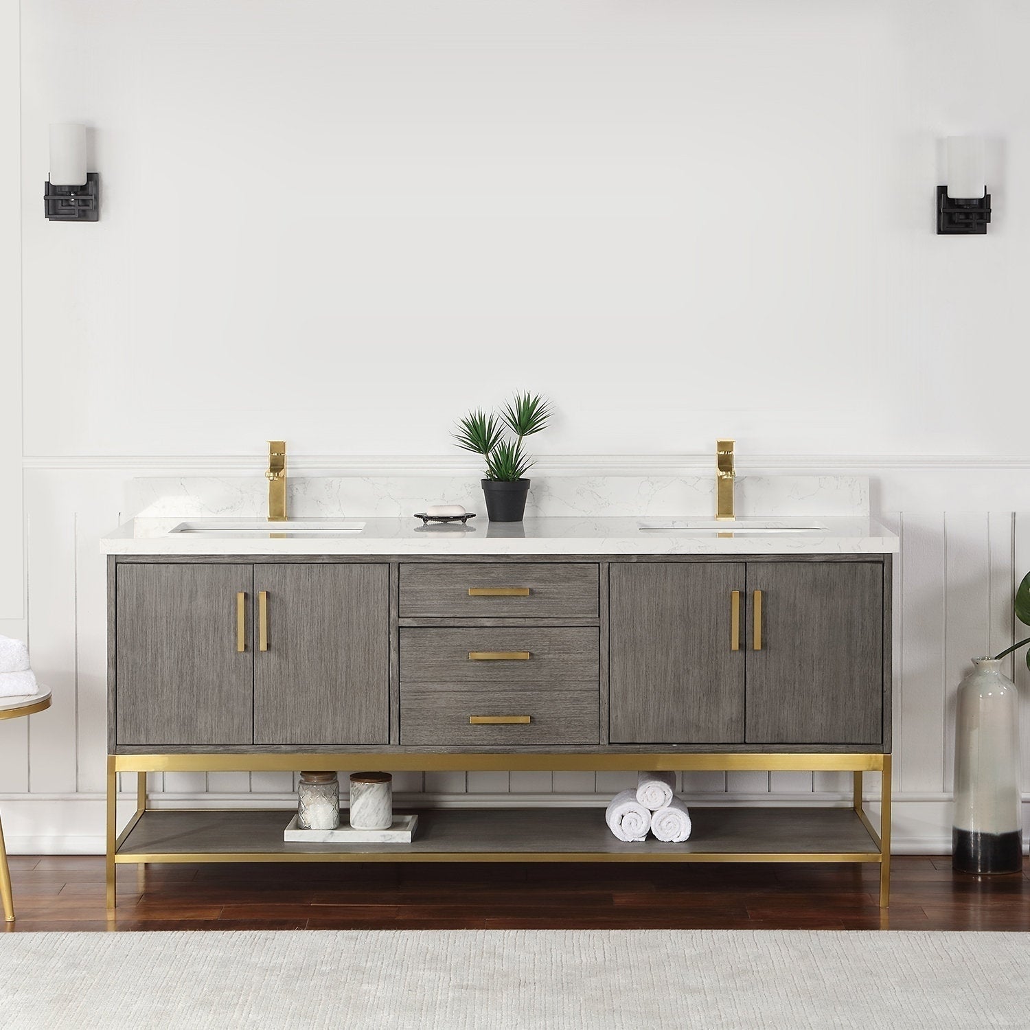 Solid wood 72-inch double-sink bathroom vanity in a stone finish with a white stone countertop, gold faucets and hardware, three center drawers, two cabinet doors and an open lower shelf with rolled towels and jars.