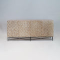 Long teak sideboard with textured checkerboard front panels in a natural finish, four doors and a slim black metal base with crisscross stretcher, photographed against a neutral studio background.