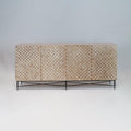 Long teak sideboard with textured checkerboard front panels in a natural finish, four doors and a slim black metal base with crisscross stretcher, photographed against a neutral studio background.