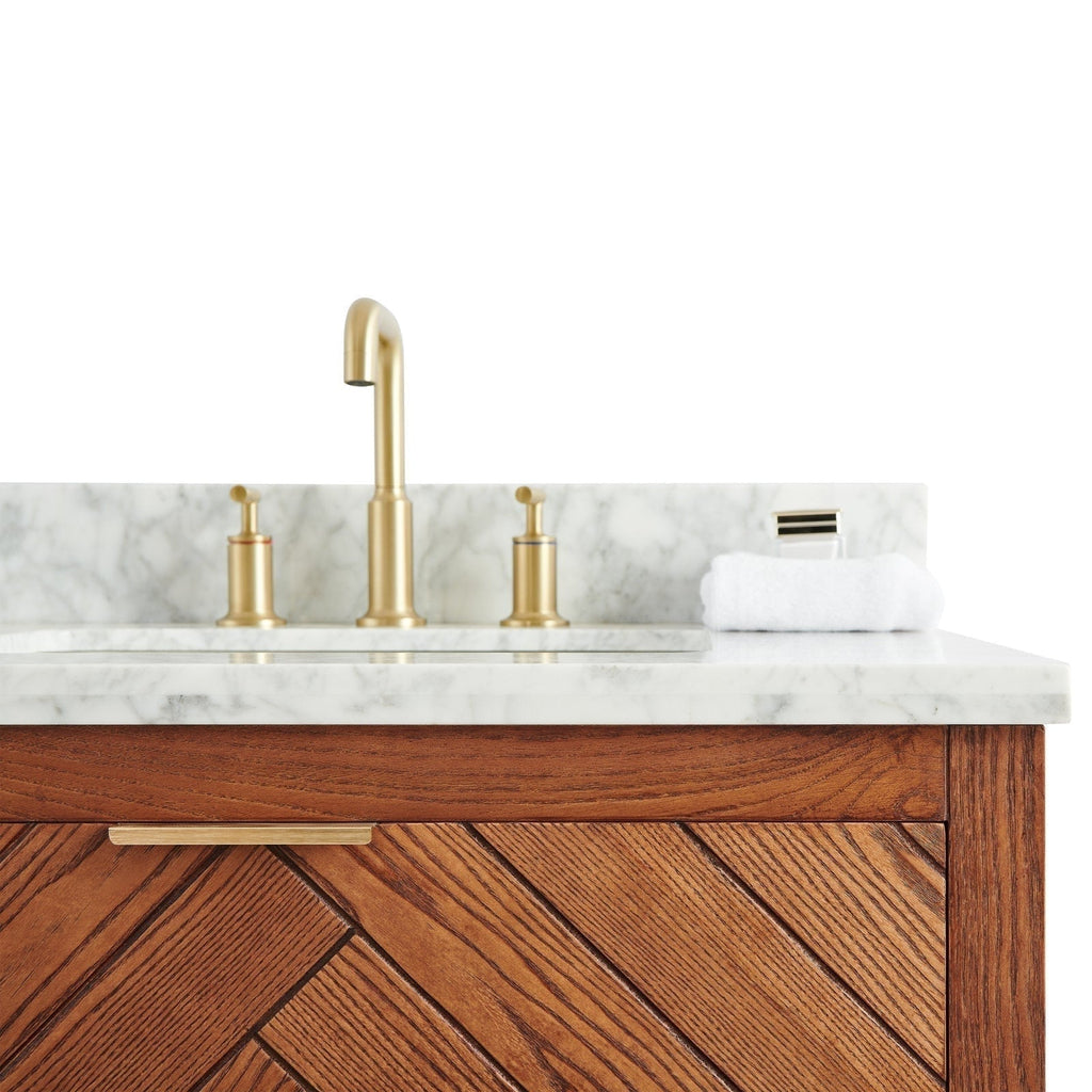 Close-up of a stone marble countertop and backsplash with brushed brass faucet and matching disc handles, a folded white towel, and a warm solid wood vanity front with diagonal chevron grain and slim brass pull.