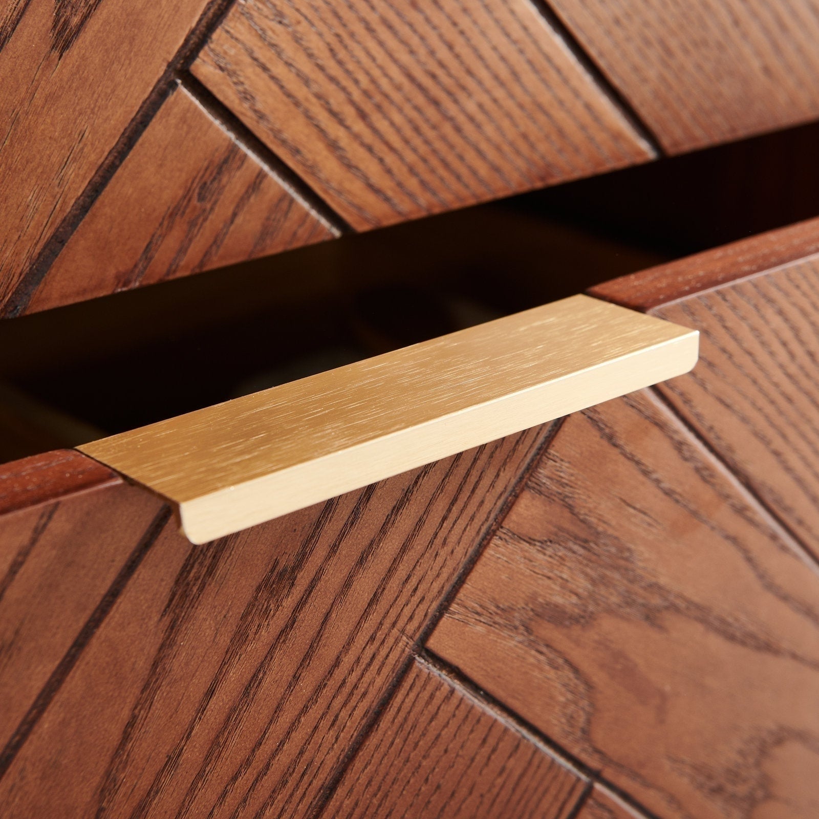 Close-up of a brushed brass rectangular drawer pull on a warm brown solid wood vanity drawer with diagonal chevron grain, drawer slightly open, showing a modern contemporary bathroom vanity detail.