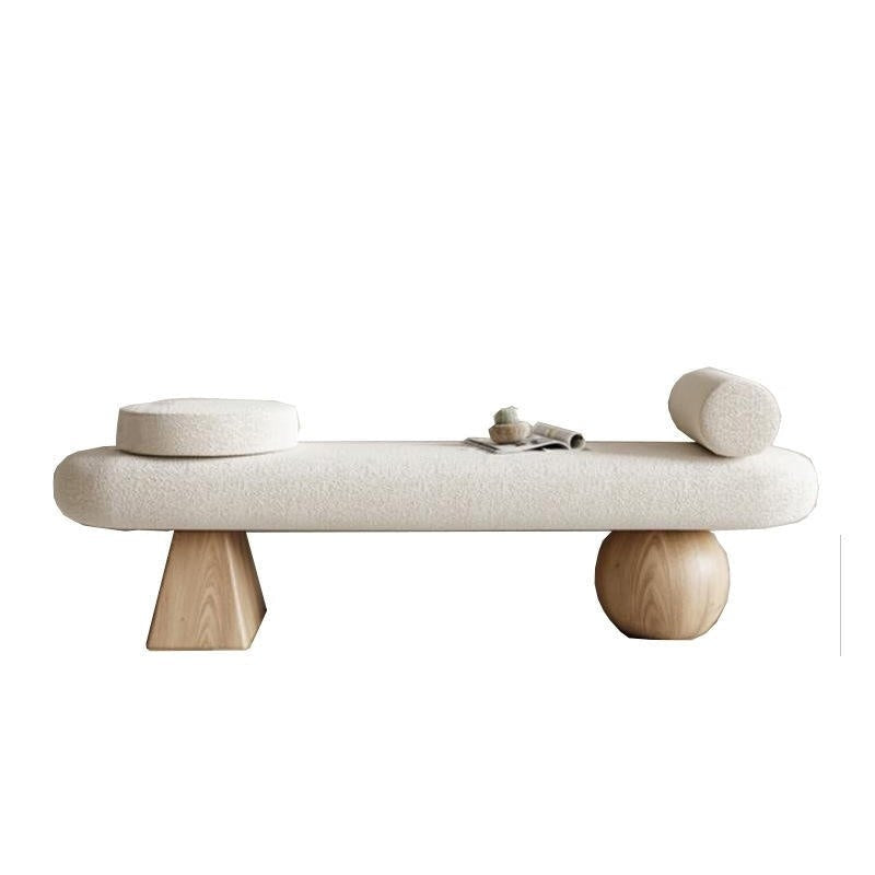 Backless cream boucle upholstered end-of-bed bench with natural wood legs, one faceted block-shaped leg and one spherical base, topped with a round disc cushion, a cylindrical bolster, and a small decorative tray.