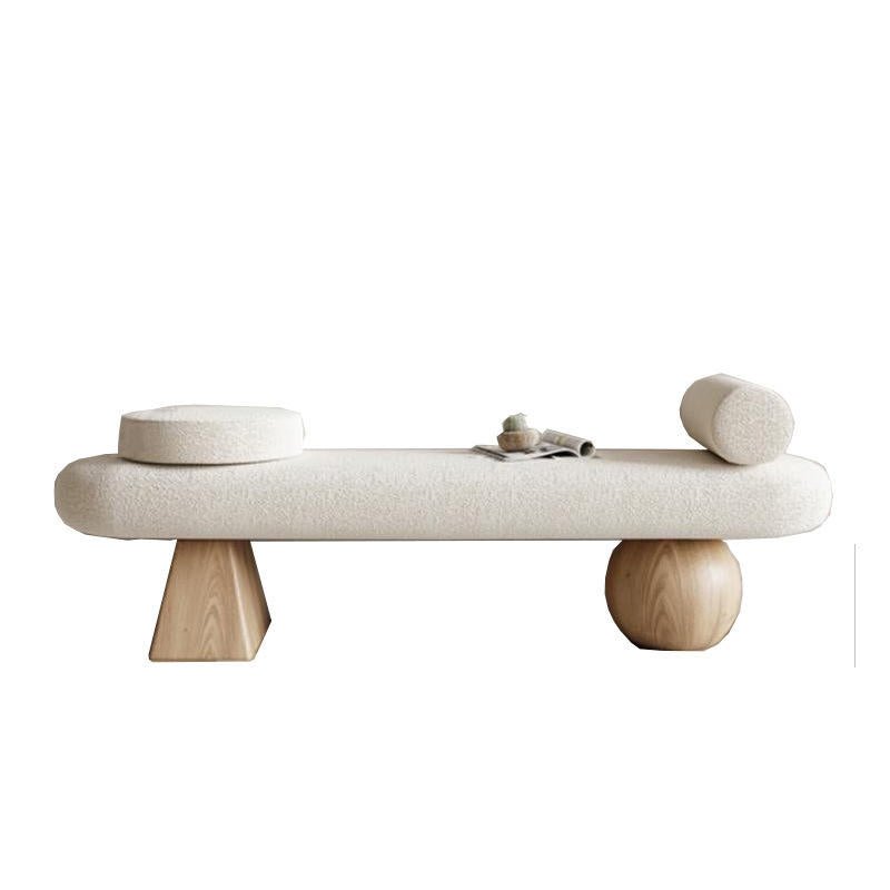 Backless cream boucle upholstered end-of-bed bench with natural wood legs, one faceted block-shaped leg and one spherical base, topped with a round disc cushion, a cylindrical bolster, and a small decorative tray.