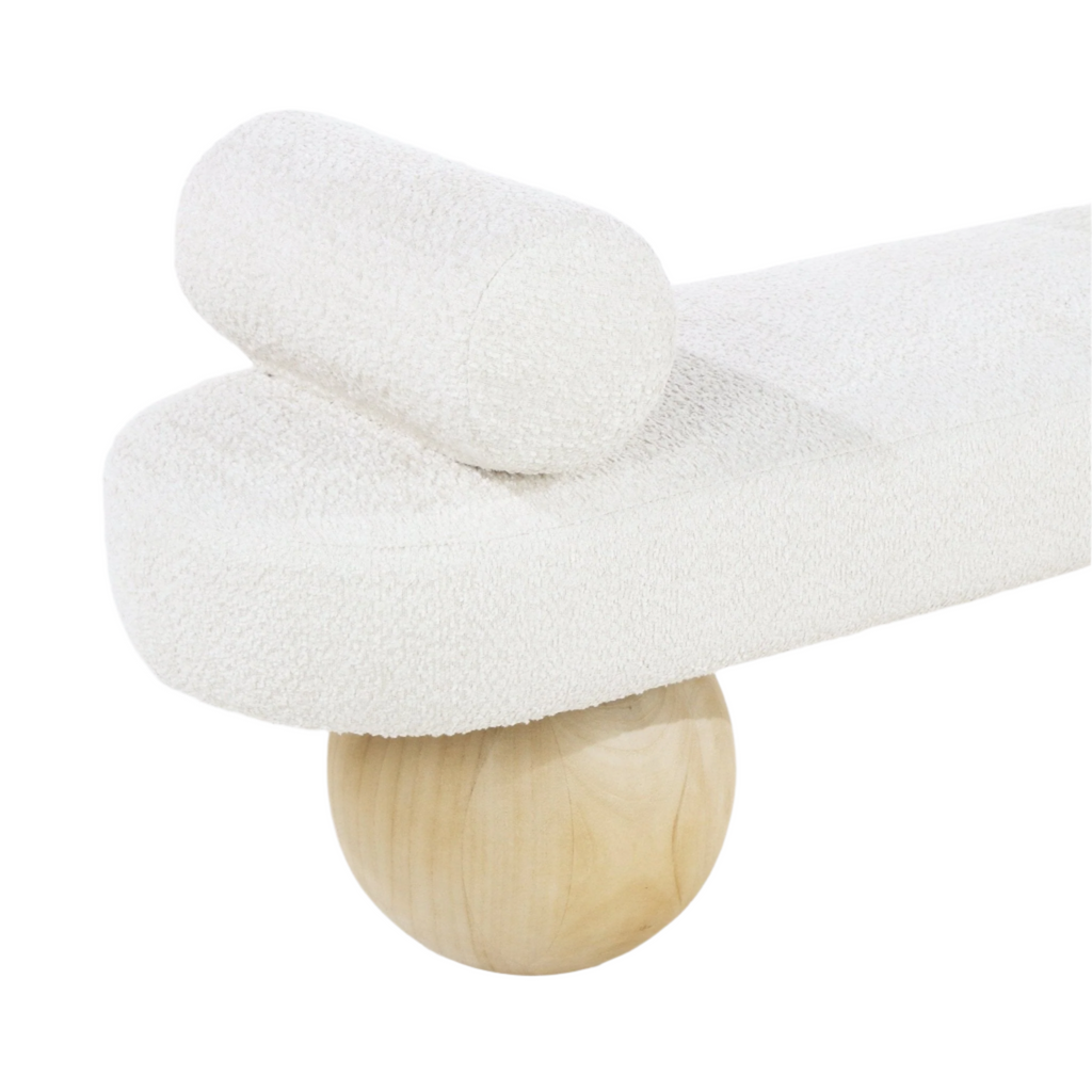 Close-up of a white boucle upholstered end-of-bed bench showing a rounded seat and cylindrical bolster perched on a spherical natural wood leg, highlighting coastal-contemporary fabric and wood materials.