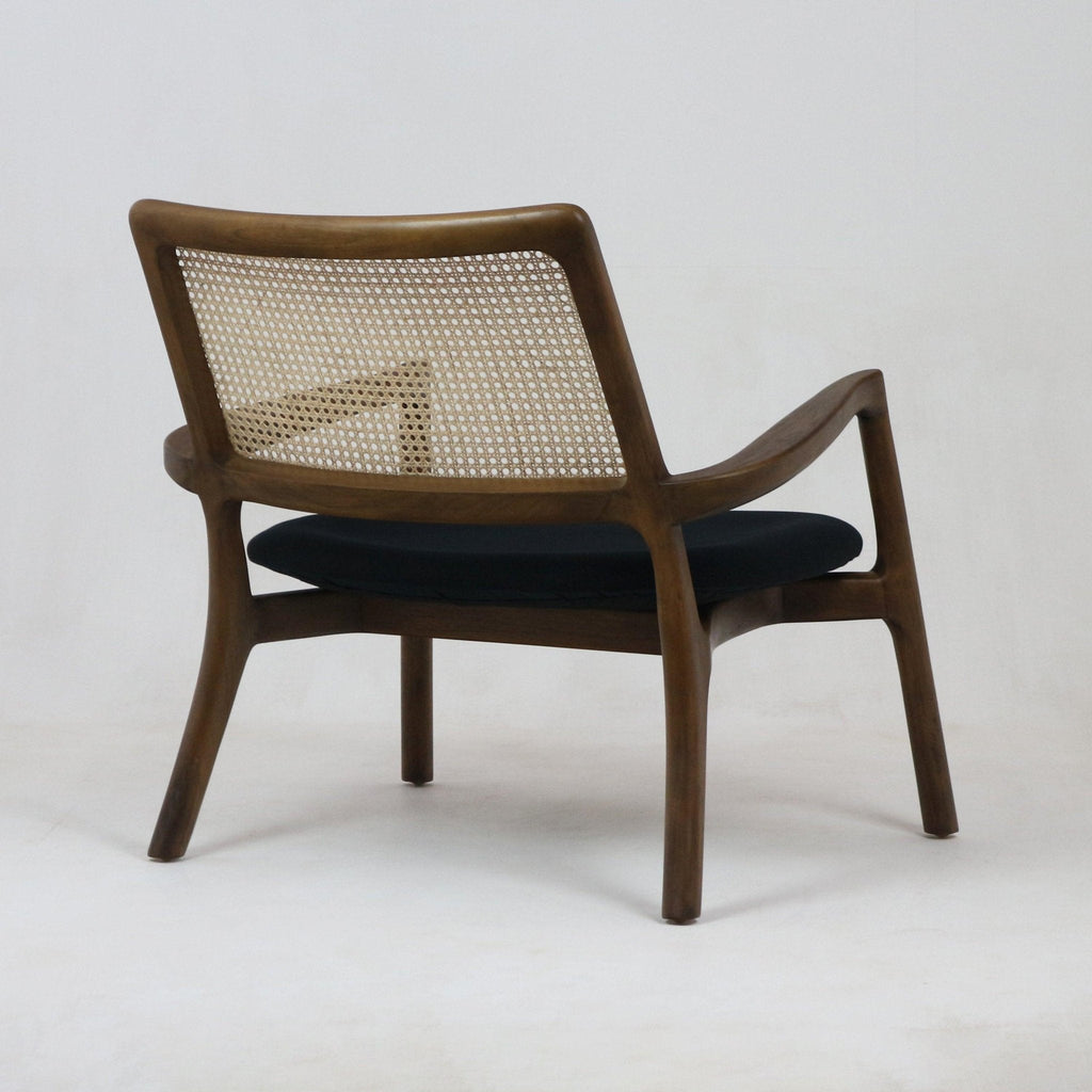 Rear three-quarter view of a mid-century modern teak lounge chair featuring a woven cane back, sculpted teak frame with curved armrests and a dark fabric seat cushion.